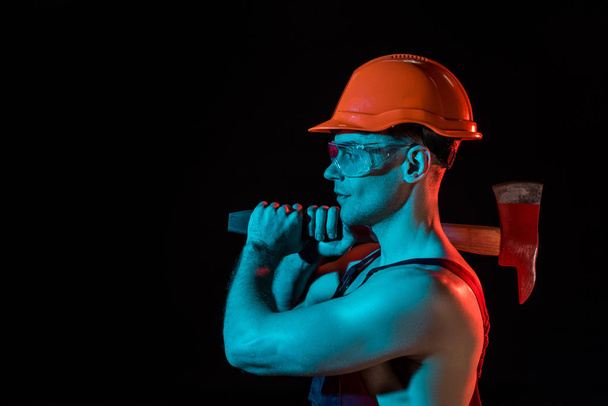 side view of sexy fireman in hardhat and protective goggles holding flat head axe isolated on black - Foto, Imagen