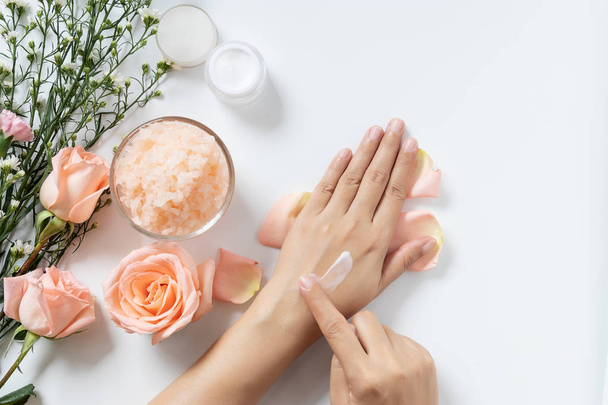 natural skincare concept. woman apply white cream on her hands on white background with jar of cosmetic cream, salt spa scrub ,rose and  white flowers with copy space - Foto, immagini