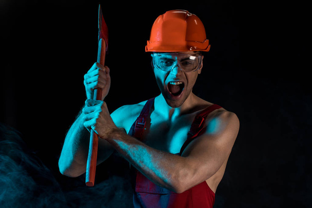 worried shirtless fireman in hardhat and protective goggles holding flat head axe and screaming in smoke on black - Zdjęcie, obraz