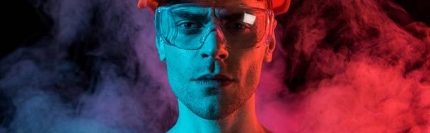 panoramic shot of serious fireman in hardhat and protective goggles in red smoke on black - Фото, изображение
