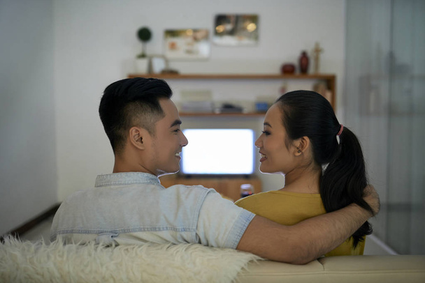 jong Vietnamees paar in liefde kijken naar elkaar wanneer zitten in de voorkant van TV set in woonkamer - Foto, afbeelding
