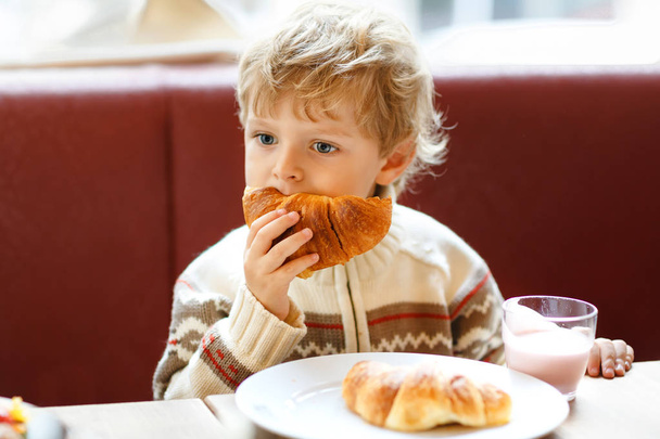 Söpö, terve poika syö croissanttia ja juo mansikkapirtelöä kahvilassa. Onnellinen lapsi aamiaisella vanhempien kanssa tai lastenhuoneessa. Vihannekset, munat terveellisenä ruokana lapsille
. - Valokuva, kuva