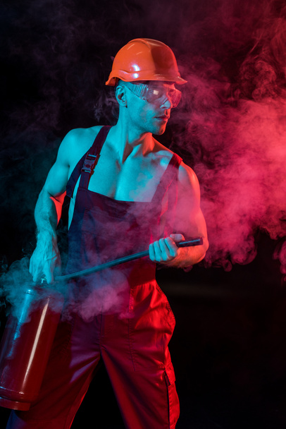 sexy hemdloser Feuerwehrmann im Overall hält Feuerlöscher in Rauch auf schwarz - Foto, Bild