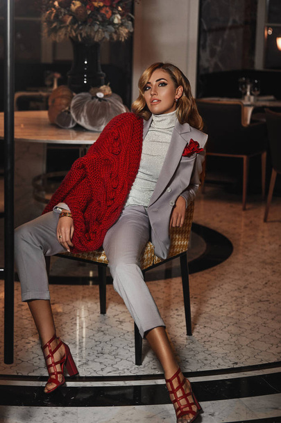 Shooting for the fashionable magazine, new autumn collection. Model wearing grey suit with high heel shoes, warm, red, knitted cardigan. Sitting on the coffee table in a lobby. Makeup and accessories. - Фото, зображення