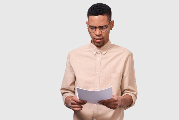 Studio portrait of serious African American young businessman in casual shirt analyzing financial reports posing on white studio background. Male entrepreneur checking paper accounting documents. - Foto, imagen