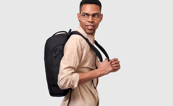 Beltéri kép fiatal afro ember hallgató hátizsák álló fehér falon. Fiatal üzletember visel alkalmi bézs inget és kerek szemüveg látszó el üres helyet a szövegben. - Fotó, kép