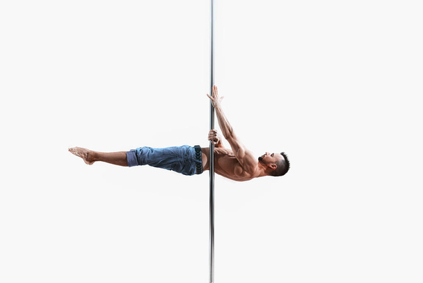 Young pole dance athlete does trick on the white background. Muscular white male sportsman practicing pole dancing.  - Photo, Image