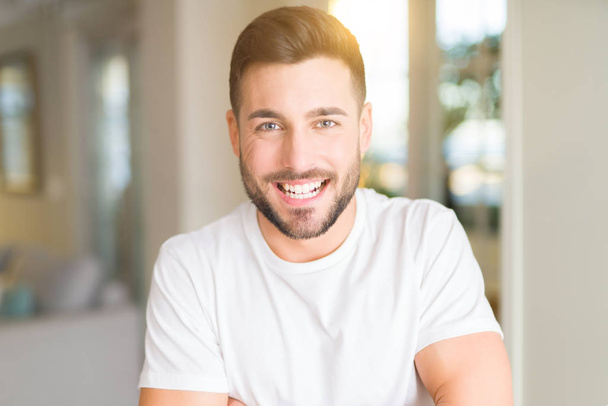 Mladý pohledný muž nosí ležérní bílé tričko doma šťastný obličej s úsměvem překřížené paže při pohledu na fotoaparát. Pozitivní člověk. - Fotografie, Obrázek