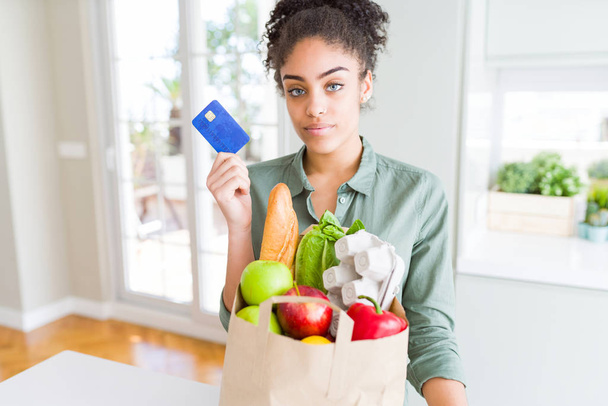 Młoda Afrykańska amerykańska dziewczyna trzyma papierową torebkę artykułów spożywczych i karty kredytowej jako płatność z pewnym wyrażeniem na inteligentne myślenie twarzy poważne - Zdjęcie, obraz