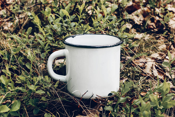 Nahaufnahme eines Metallbechers auf Waldboden zwischen Heidelbeersträuchern und Eichenblättern. Tee im Freien, Kaffeezeit. Attrappe der weißen Emaille-Tasse. Lifestyle Relax, Trekking und Campingkonzept. - Foto, Bild