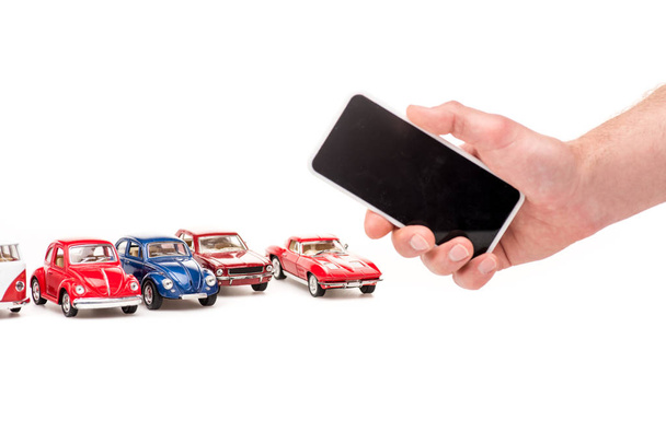 bijgesneden beeld van man houden smartphone met blanco scherm en kleurrijke speelgoed auto's op wit - Foto, afbeelding