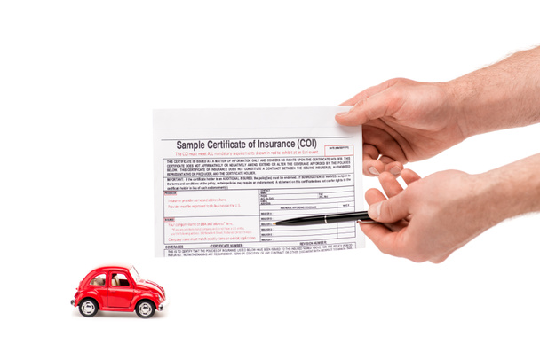 bijgesneden weergave van de man met pen en verzekeringscertificaat in de buurt van rode speelgoed auto geïsoleerd op wit - Foto, afbeelding