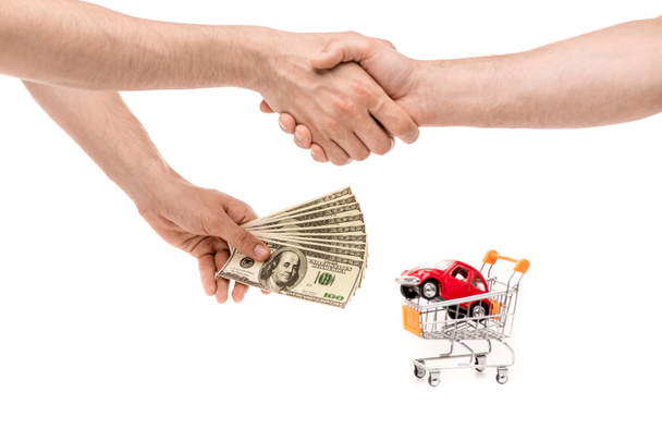 cropped view of men with dollar banknotes shaking hands near toy car in shopping cart isolated on white - Photo, Image