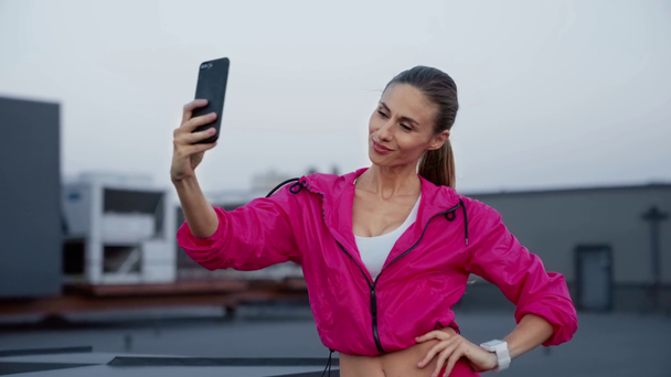 charming sportswoman in sportswear holding smartphone in hands, posing and taking selfie, standing on rooftop - Footage, Video