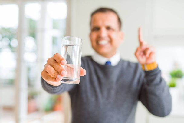 Homem de meia-idade bebendo copo de água em casa surpreso com uma ideia ou pergunta apontando o dedo com o rosto feliz, número um
 - Foto, Imagem