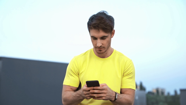 sarı spor giyim yakışıklı sporcu çatıda oturan, elinde akıllı telefon tutarak, mesaj mesajlaşma, gülümseyerek ve ciddi yüz yapma - Video, Çekim