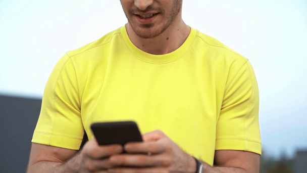 low angle view of sportsman in yellow sportswear sitting on rooftop, holding smartphone in hands, texting message, smiling and making serious face - Video, Çekim