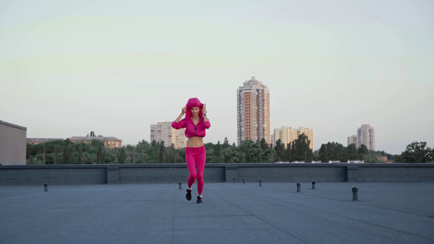 happy sportswoman running on rooftop and putting on hood of sports sweater on head - Video, Çekim