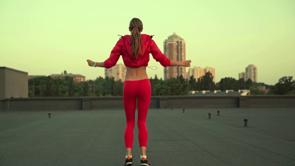 beautiful sportswoman in sportswear holding hands over head, jumping  in place and side to side, standing on rooftop - Footage, Video