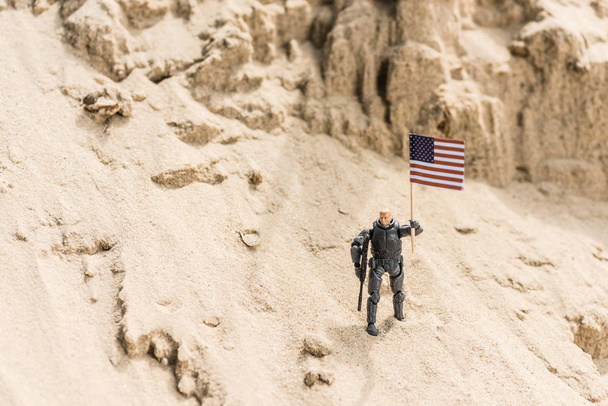 おもちゃの武装兵士が砂の上に立ち、アメリカ国旗を掲げている - 写真・画像