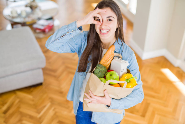Красивая молодая женщина держит бумажный пакет, полный здоровых продуктов с счастливым лицом улыбается делает хорошо знак с рукой на глазу, глядя сквозь пальцы
 - Фото, изображение