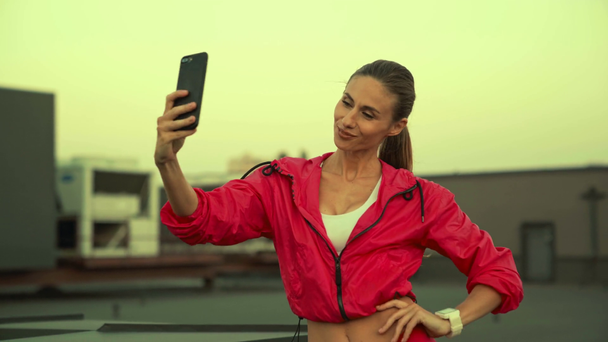 bela esportista em sportswear rosa segurando smartphone em mãos, posando e tirando selfie, de pé no telhado
 - Filmagem, Vídeo