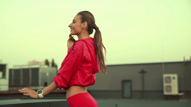 щаслива спортсменка в рожевому спортивному одязі тримає смартфон в руках і розмовляє, стоїть на даху
 - Кадри, відео