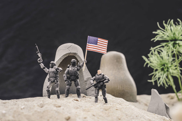 selective focus of toy soldiers with guns and american flag standing near caves on sand dune  - 写真・画像