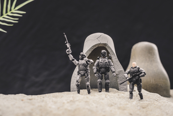 speelgoed soldaten met geweren en Amerikaanse vlag staande in de buurt van grotten op zandduin op zwarte achtergrond - Foto, afbeelding
