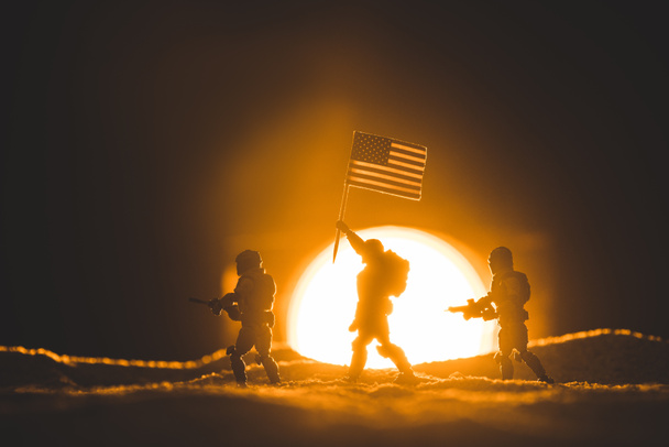 selective focus of toy soldiers silhouettes with guns and american flag walking on planet with sun on background - Photo, Image