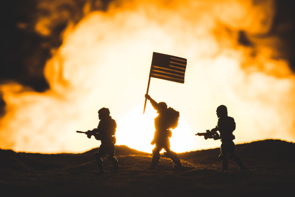 παιχνίδια στρατιώτες φιγούρες με όπλα και αμερικάνικη σημαία περπατώντας στον πλανήτη με ήλιο σε καπνό στο παρασκήνιο - Φωτογραφία, εικόνα