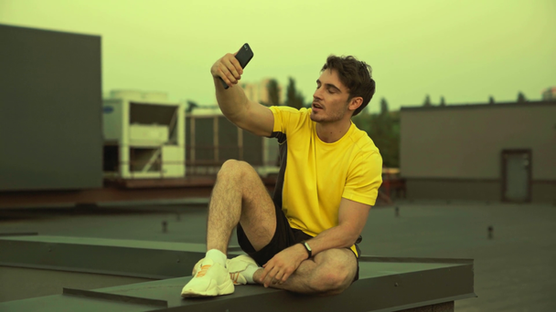 handsome sportsman sitting on rooftop, holding smartphone in hands, smiling, making grimace and showing victory sign, taking selfie - Footage, Video