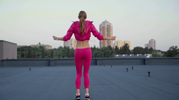 charming sportswoman in sportswear holding hands over head, jumping in place and side to side, standing on rooftop - Footage, Video