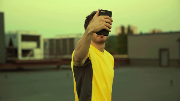 handsome sportsman in yellow sportswear holding smartphone in hand, talking on video chat, showing victory sign and standing on rooftop - Footage, Video