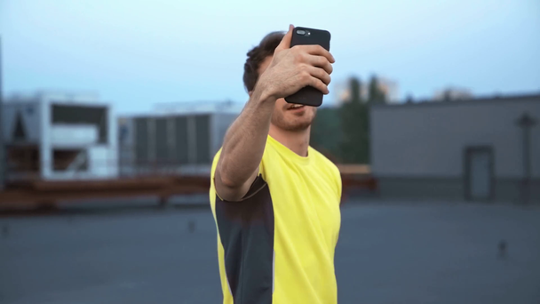 schöner Sportler in gelber Sportbekleidung hält Smartphone in der Hand, spricht im Videochat, zeigt Siegeszeichen und steht auf dem Dach - Filmmaterial, Video