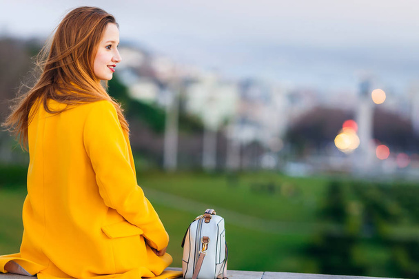 спина девушки в желтом пальто, сидящей на фоне города - Фото, изображение