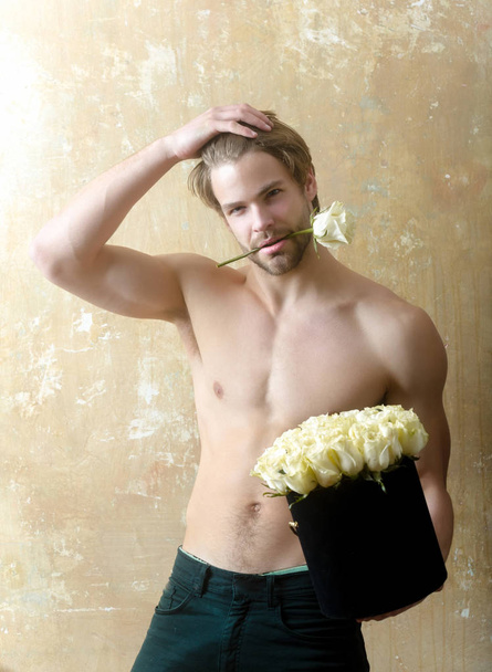 Ο άνθρωπος με το μπουκέτο τριαντάφυλλα. Ο σέξι μισόγυμνος όμορφος τύπος αγγίζει τα μαλλιά. Ωραίο σώμα και γοητευτικά λουλούδια. Σαγηνευτικό ποζάρω με λουλούδι στα δόντια - Φωτογραφία, εικόνα