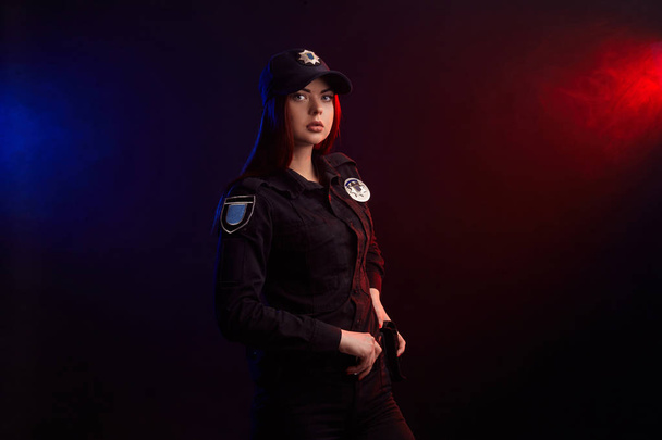 Mujer policía seria posando para la cámara sobre un fondo negro con retroiluminación roja y azul
. - Foto, imagen