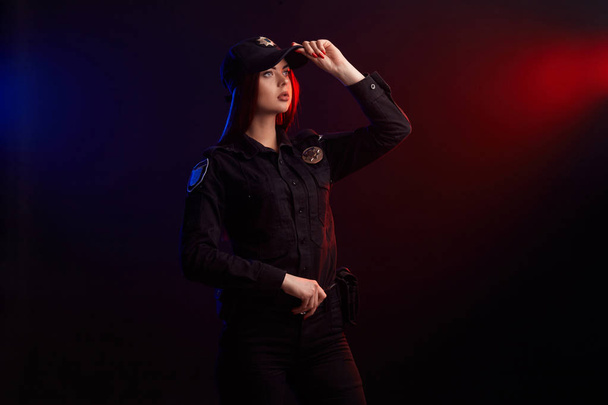 Vakava naispoliisi poseeraa kameralle mustaa taustaa vasten punaisella ja sinisellä taustavalolla
. - Valokuva, kuva