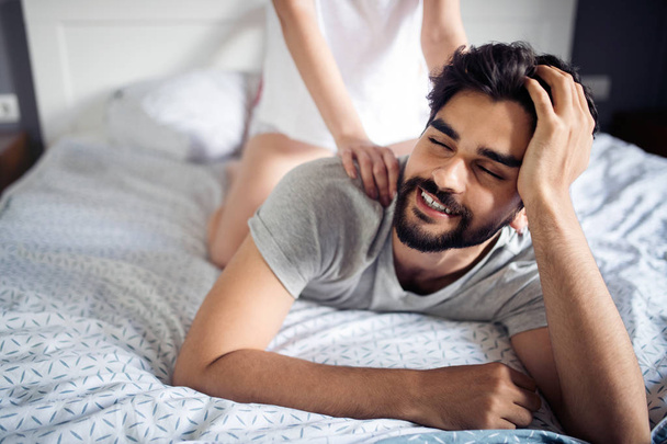 Beautiful young woman is doing her handsome boyfriend massage and smiling while they are resting at home - Photo, Image
