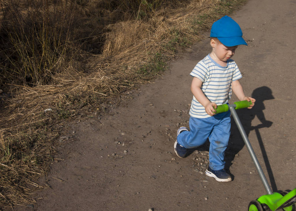 Wczesna wiosna, mały chłopiec z trzech lat w niebieskich spodniach z zieloną skuter na drodze wiejskiej - Zdjęcie, obraz