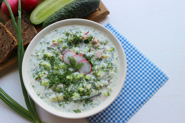 Okroshka is a traditional Russian summer cold soup in a ceramic bowl. Ingredients potatoes, radishes, cucumbers, dill, eggs, kefir yogurt, sausage. Summer yogurt cold soup on a wooden table. Selective focus, copy space. - Fotoğraf, Görsel