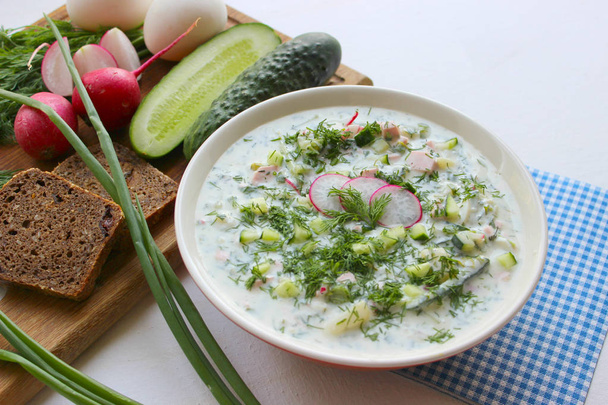 Okroshka is a traditional Russian summer cold soup in a ceramic bowl. Ingredients potatoes, radishes, cucumbers, dill, eggs, kefir yogurt, sausage. Summer yogurt cold soup on a wooden table. Selective focus, copy space. - Foto, imagen