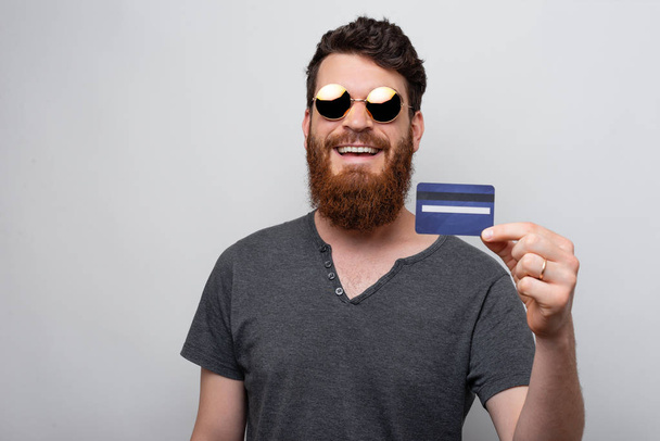 Cheerful man with beard holding blue credit card over grey background - Foto, Imagem