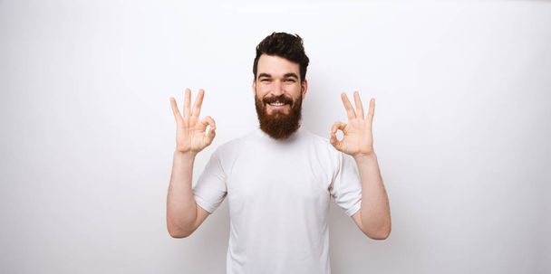 A fiatal szakállas férfi portréja, az OK gesztus fehér háttérrel - Fotó, kép