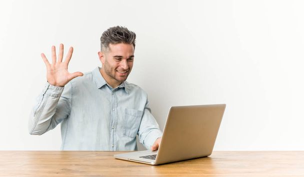junger, gutaussehender Mann, der mit seinem Laptop arbeitet und fröhlich lächelt und Nummer fünf mit den Fingern zeigt. - Foto, Bild