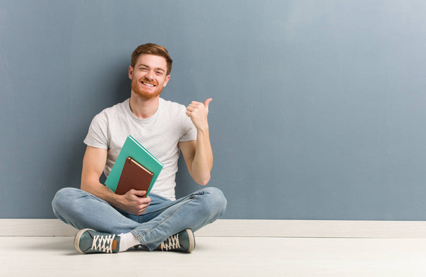 Young redhead student man sitting on the floor pointing to the side with finger. He is holding books. - Fotoğraf, Görsel