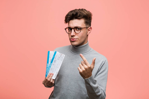 Young cool man holding an air tickets pointing with finger at you as if inviting come closer. - Foto, imagen