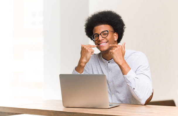 Молодой чернокожий человек с помощью ноутбука улыбается, показывая рот
 - Фото, изображение