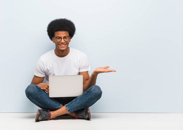 Giovane uomo nero seduto sul pavimento con un computer portatile che tiene qualcosa con le mani
 - Foto, immagini